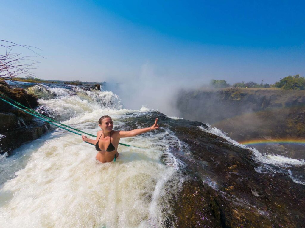 Tourists swimming at the edge of Victoria Falls in Devil’s Pool, with water rushing past and panoramic views of the gorge below