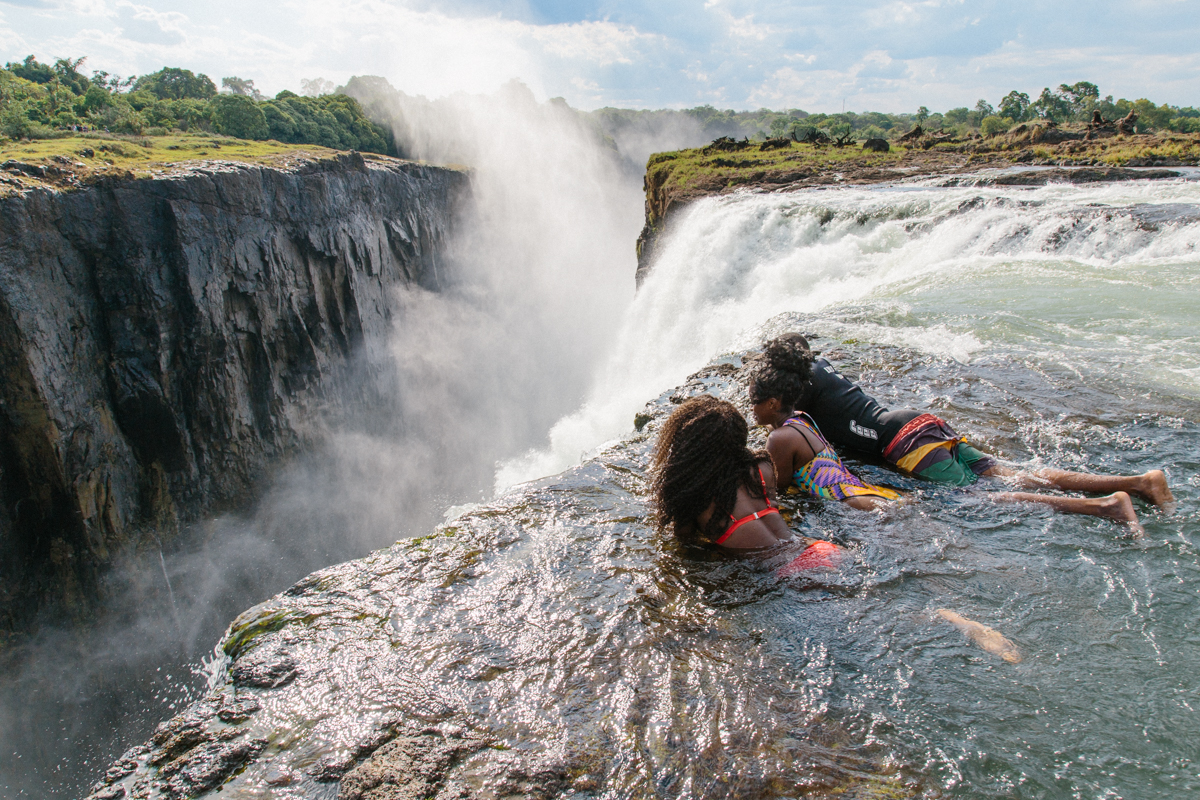 Tourists swimming at the edge of Victoria Falls in Devil’s Pool, with water rushing past and panoramic views of the gorge below