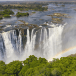 A panoramic view of Victoria Falls with cascading water and vibrant rainbow mist, showcasing the majestic beauty of the Zambezi River between Zimbabwe and Zambia.