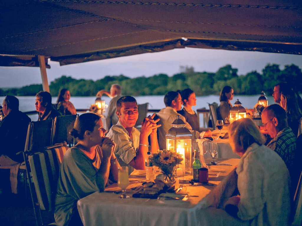 Guests enjoying a sunset dinner on a Zambezi River cruise near Victoria Falls, with candlelit tables, calm waters, and wildlife along the riverbank
