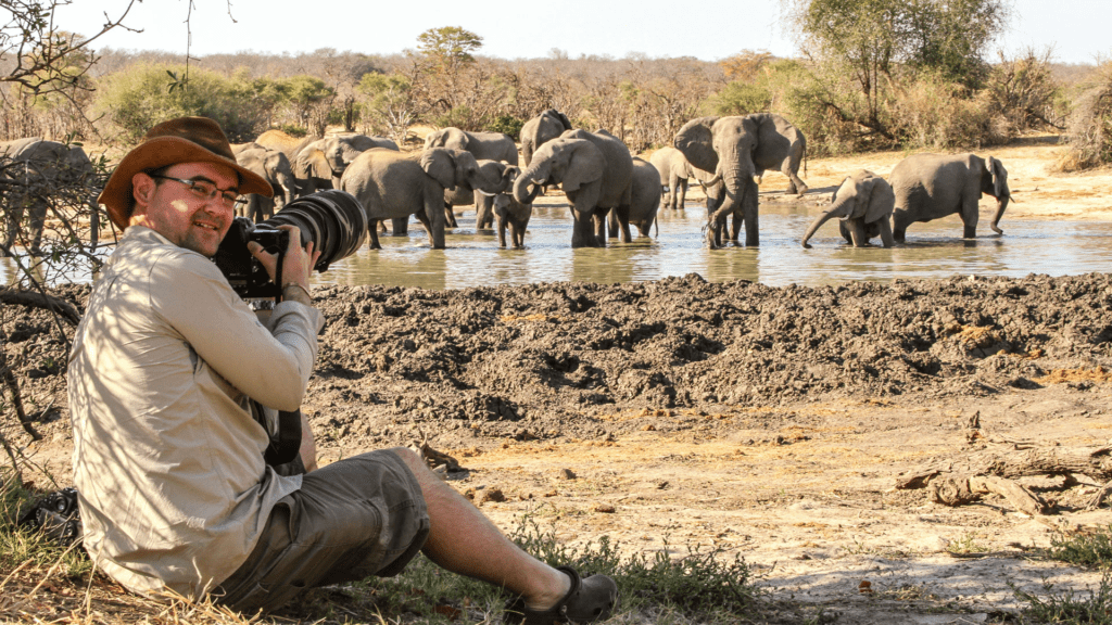 Photographer with a telephoto lens sitting near a waterhole while photographing a herd of elephants in Victoria Falls region