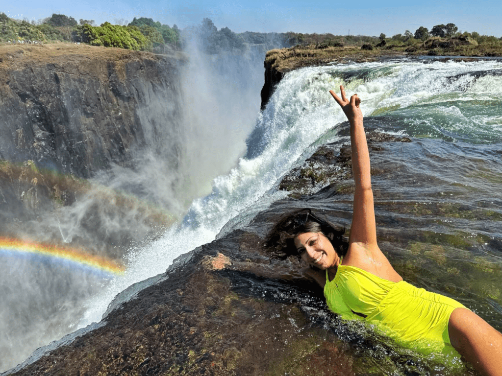 Smiling woman in yellow swimsuit enjoying the Devil’s Pool at the edge of Victoria Falls on the Zambia side, with a rainbow in the mist